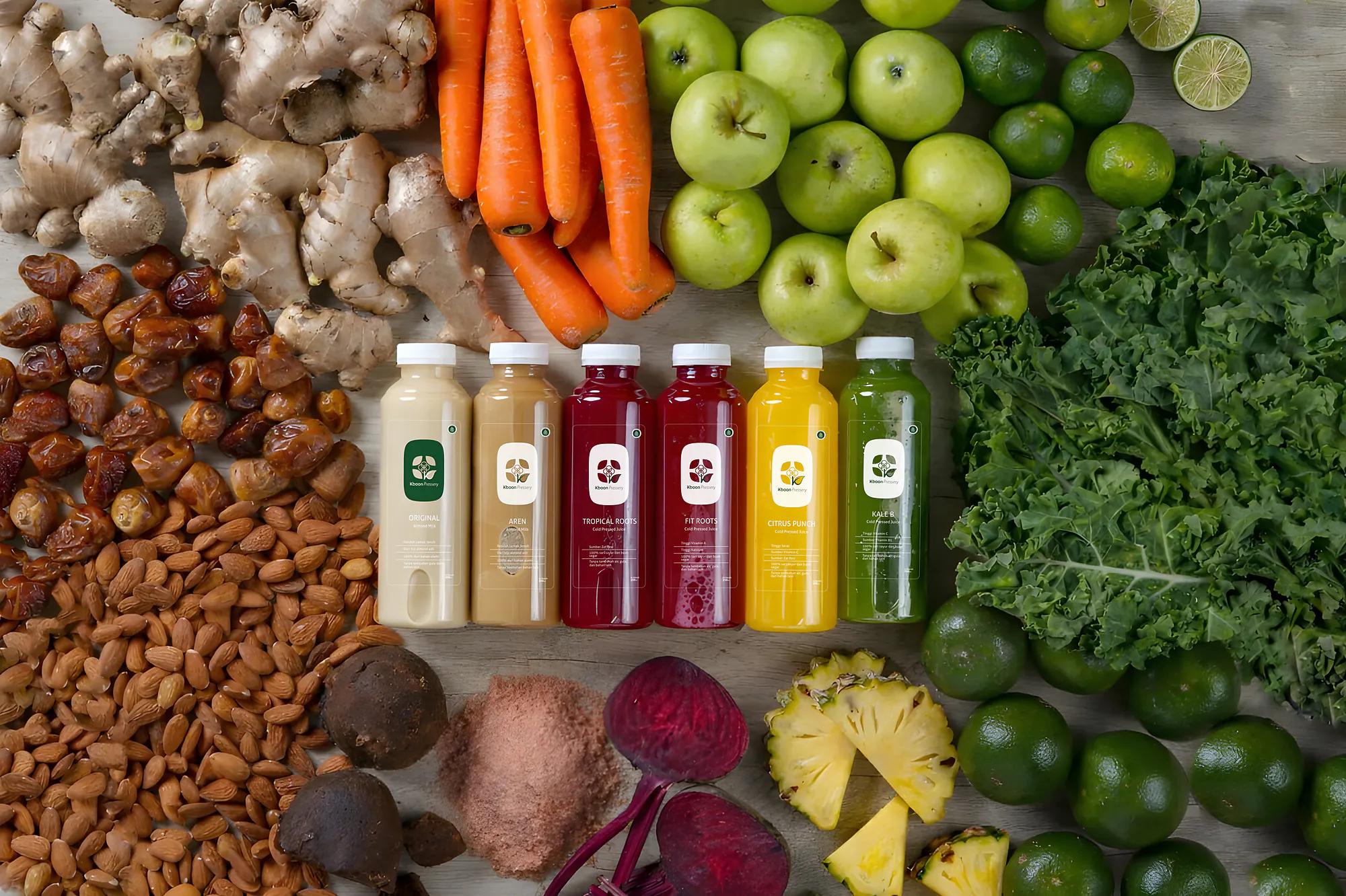 Enam botol Kboon (Original, Aren, Tropical Roots, Fit Roots, Citrus Punch, Kale B) berjajar di atas meja kayu, dikelilingi jahe, wortel, apel hijau, jeruk nipis, kale, nanas, bit, almond, dan kurma