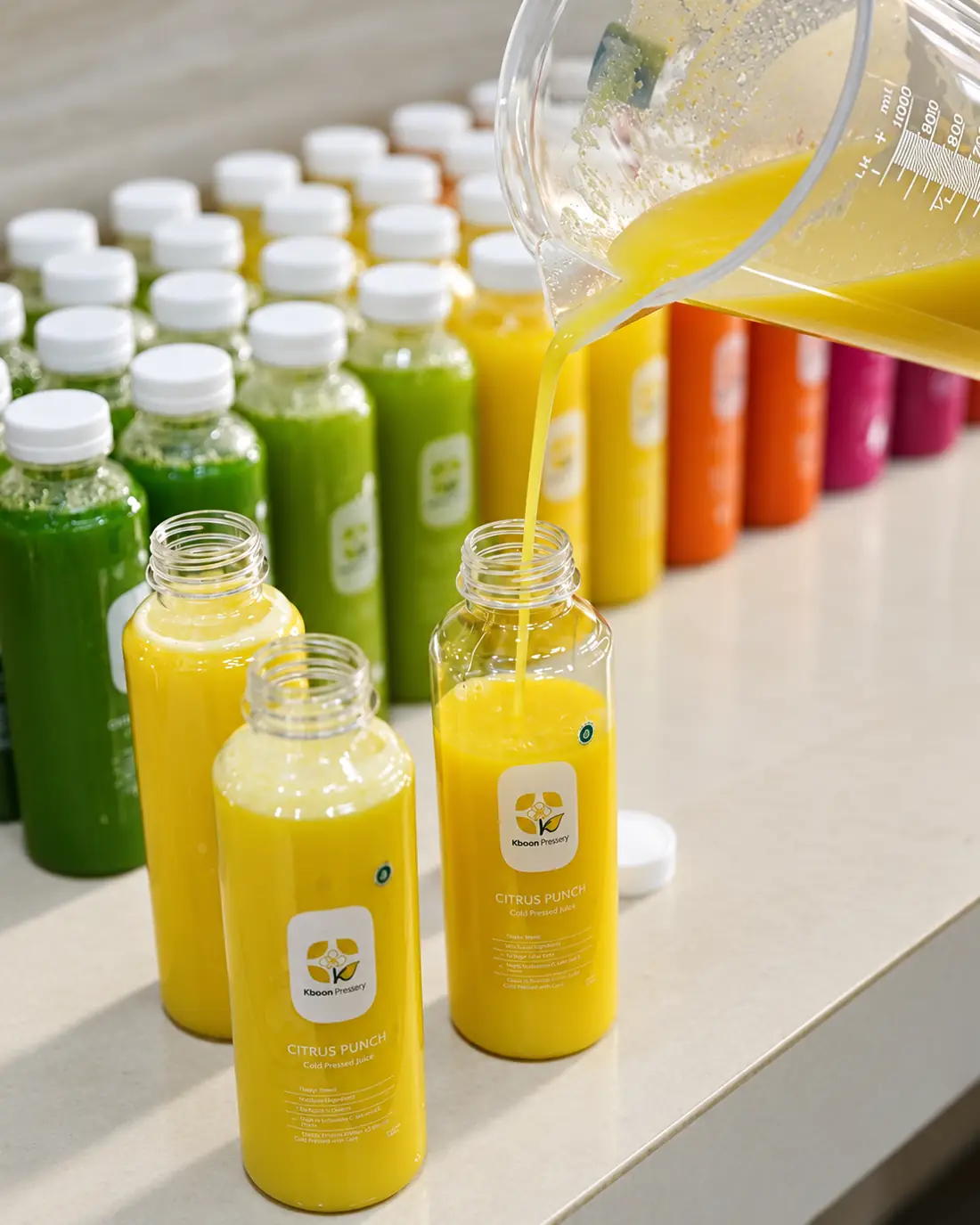 Jus kuning dituang dari teko takar ke deretan botol Kboon